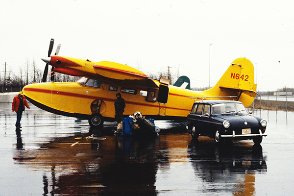 A bright yellow seaplane parked beside a vintage black car, with rainy skies above.