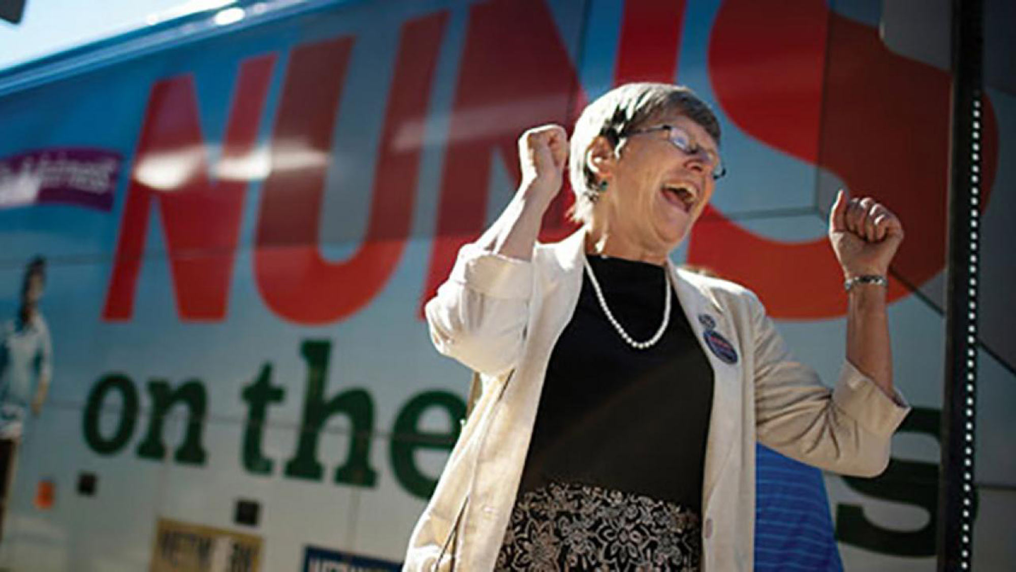 Smiling woman raises arms in triumph outside bus with sign "Nuns on the Bus"