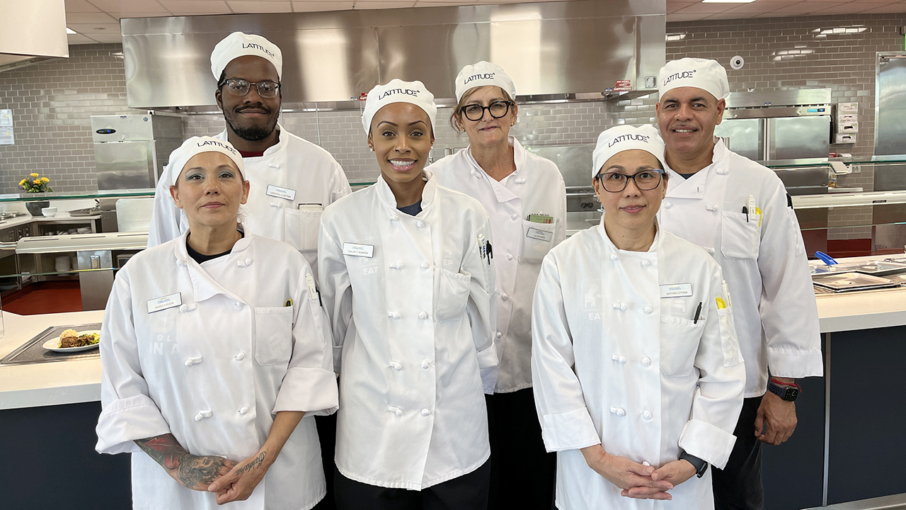Dressed in kitchen whites, the six participants in the Culinary Training Program stand in front of a serving platform at Latitude.Latitude
