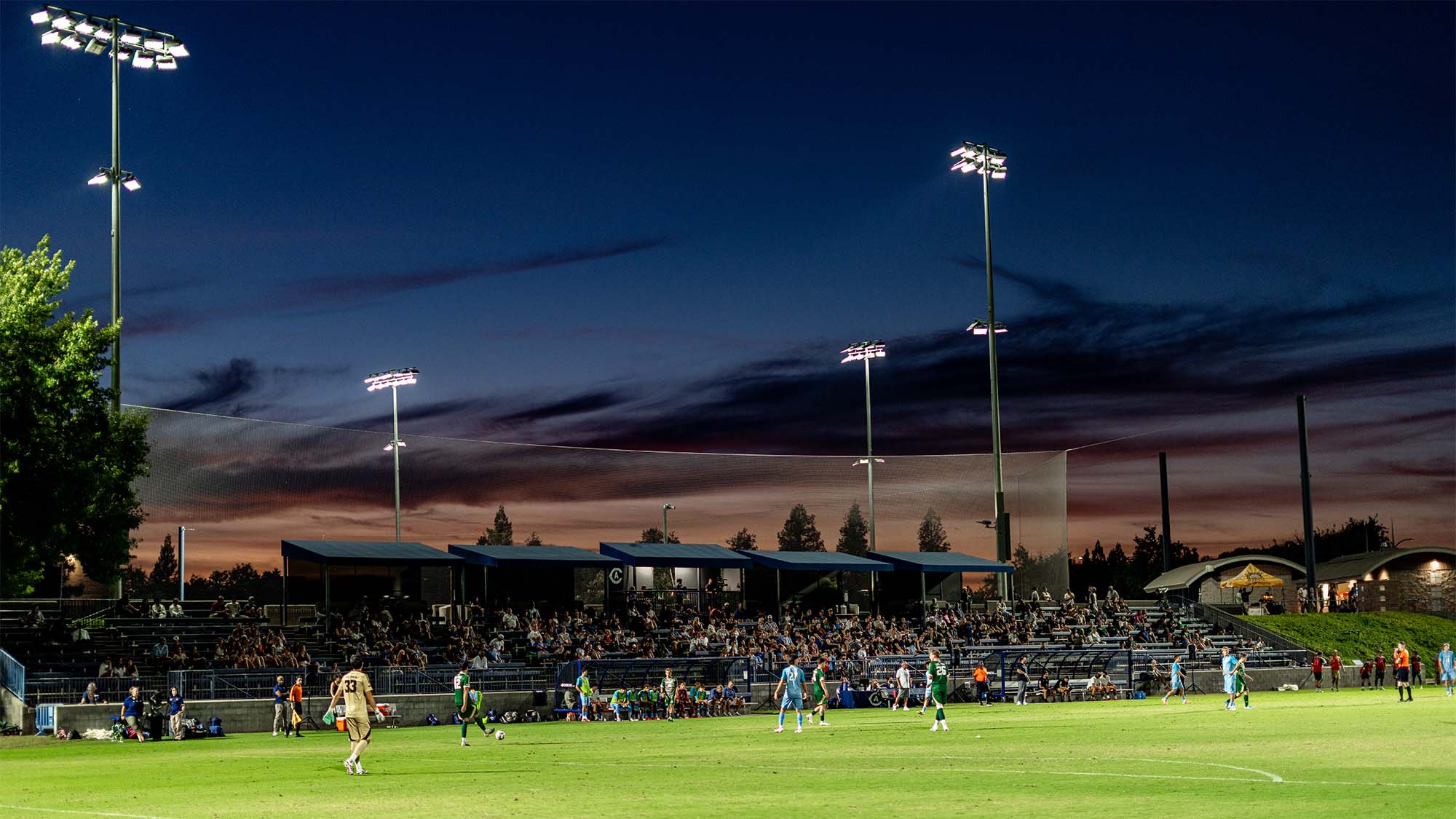 Evening soccer match with players under tall floodlights, packed stands and twilight sky
