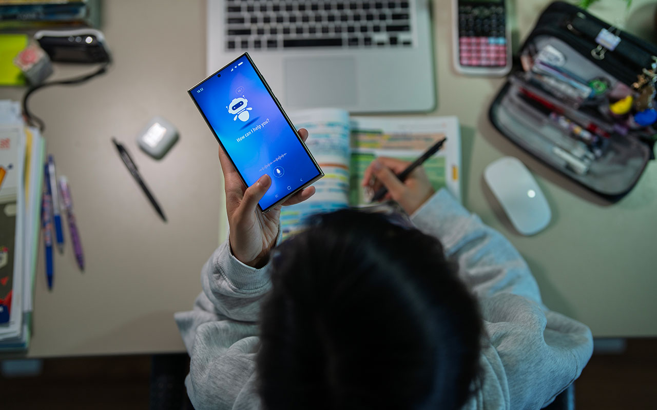 A student sits at a desk studying with an open notebook while holding a smartphone displaying an AI assistant screen.