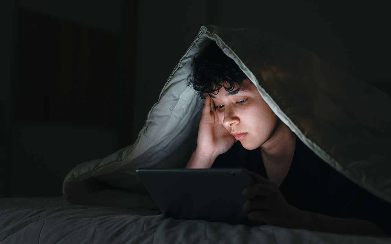 A person lying in bed at night with a blanket over their head, looking at a tablet screen that lights up their face.