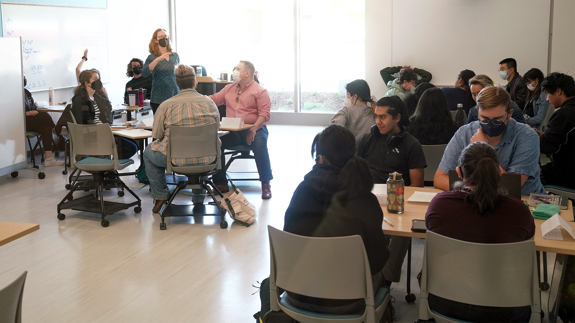 Instructor speaks while students in desks — some arranged in circles — listen.