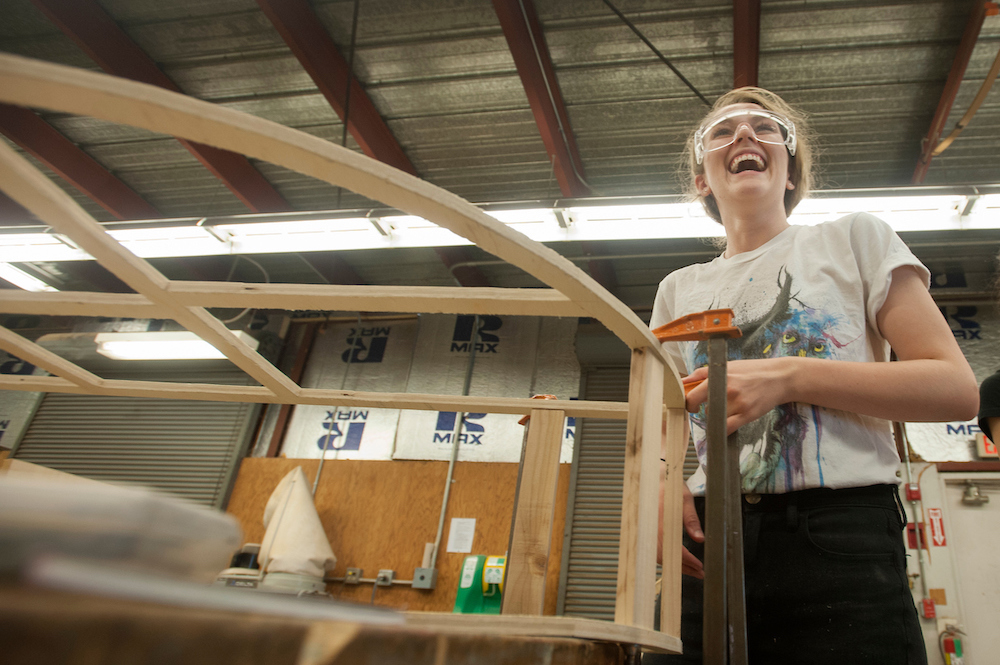 A student wearing goggles laughs beside the scaffolding of her furniture at UC Davis.