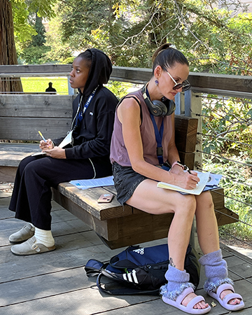 Two students write as they are seated on a bench on the deck of the Wyatt Pavilion in the Arboretum