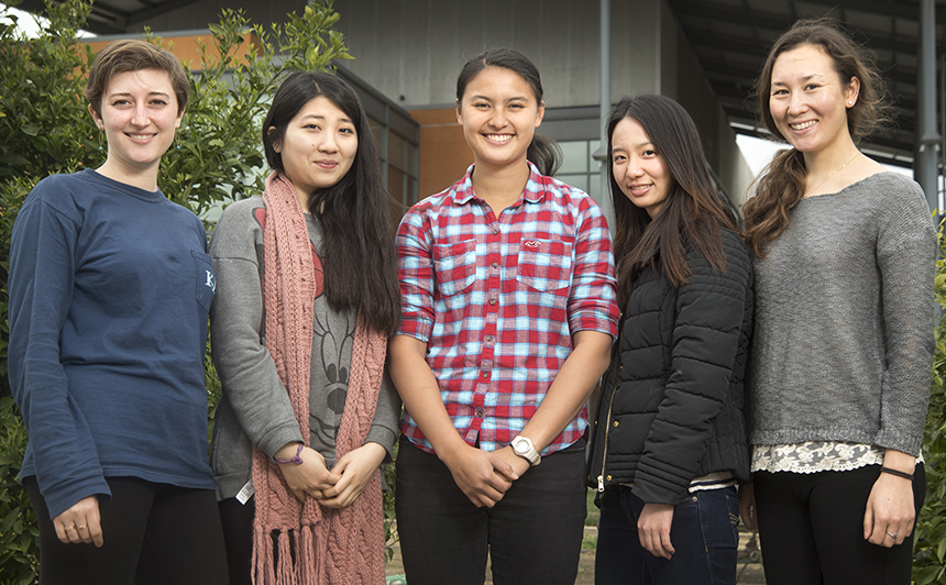 Five college women stand in garden in front of two-story orange building.