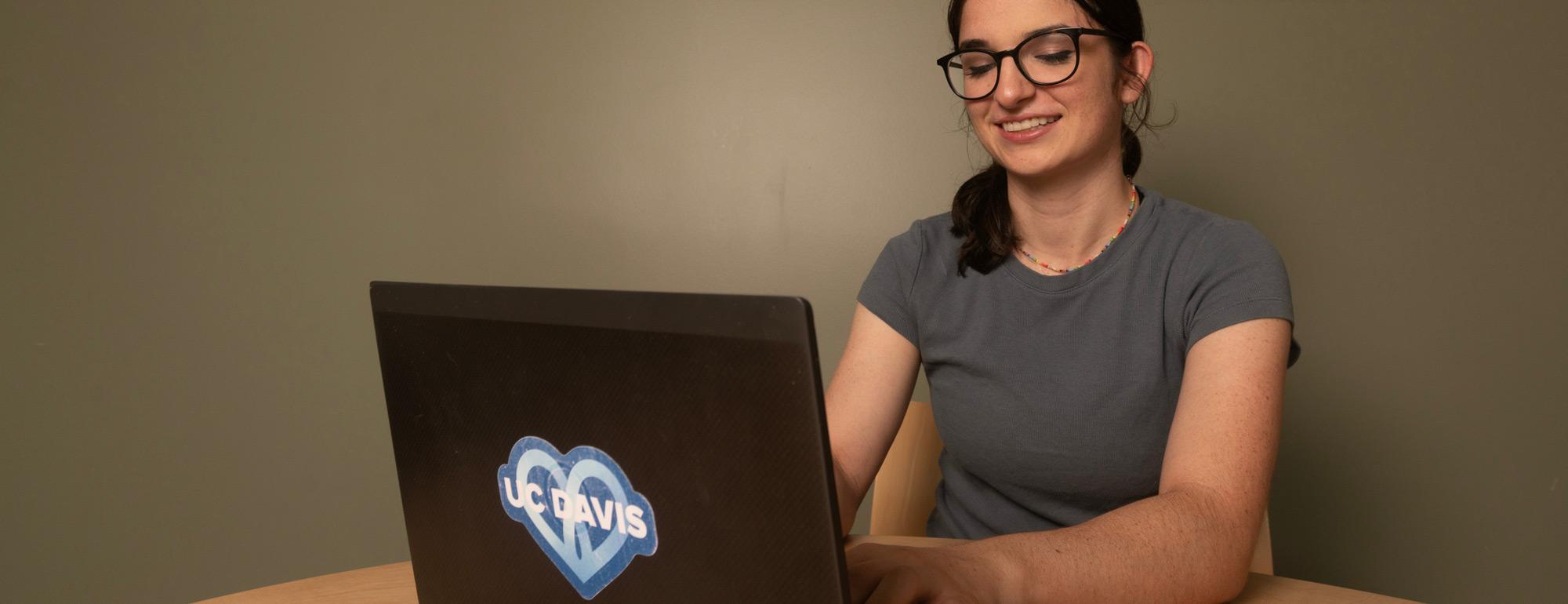 Smiling woman with glasses seated at a laptop displaying a heart sticker