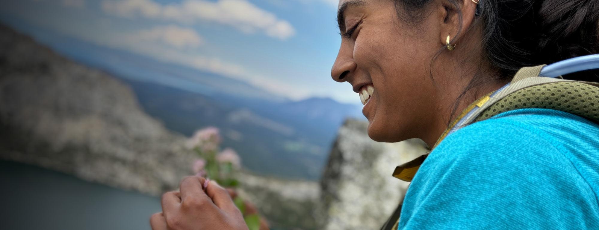 Smiling woman in blue jacket holding wildflower with mountains and lake behind
