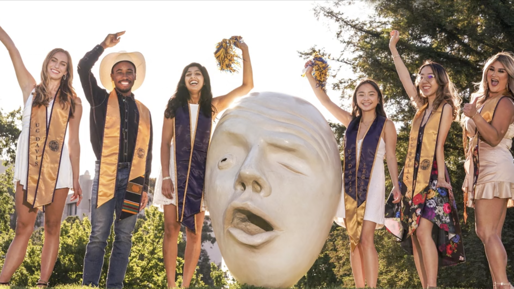 six students in graduation hats and sashes pose near an Egghead statue at UC Davis