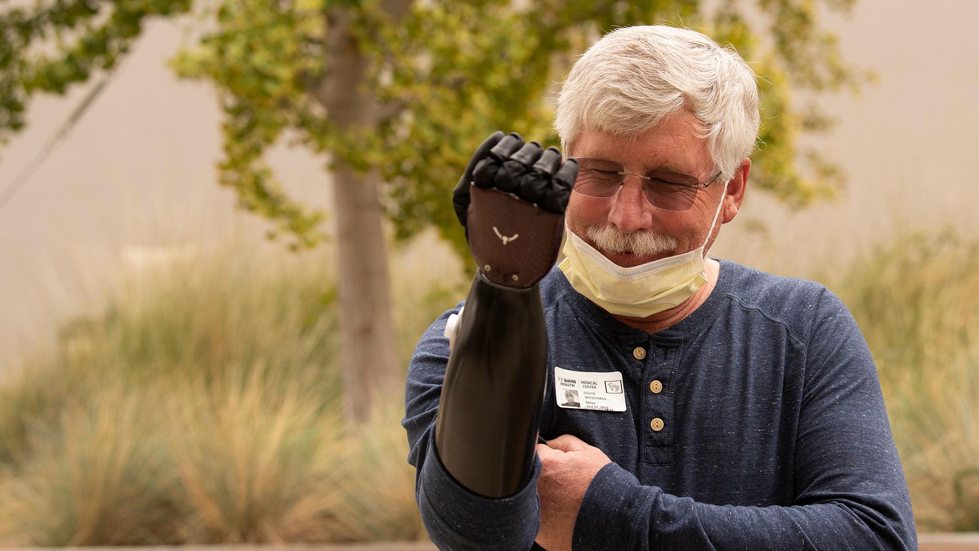 Older man outdoors smiling, wearing a black prosthetic arm and a lowered face mask