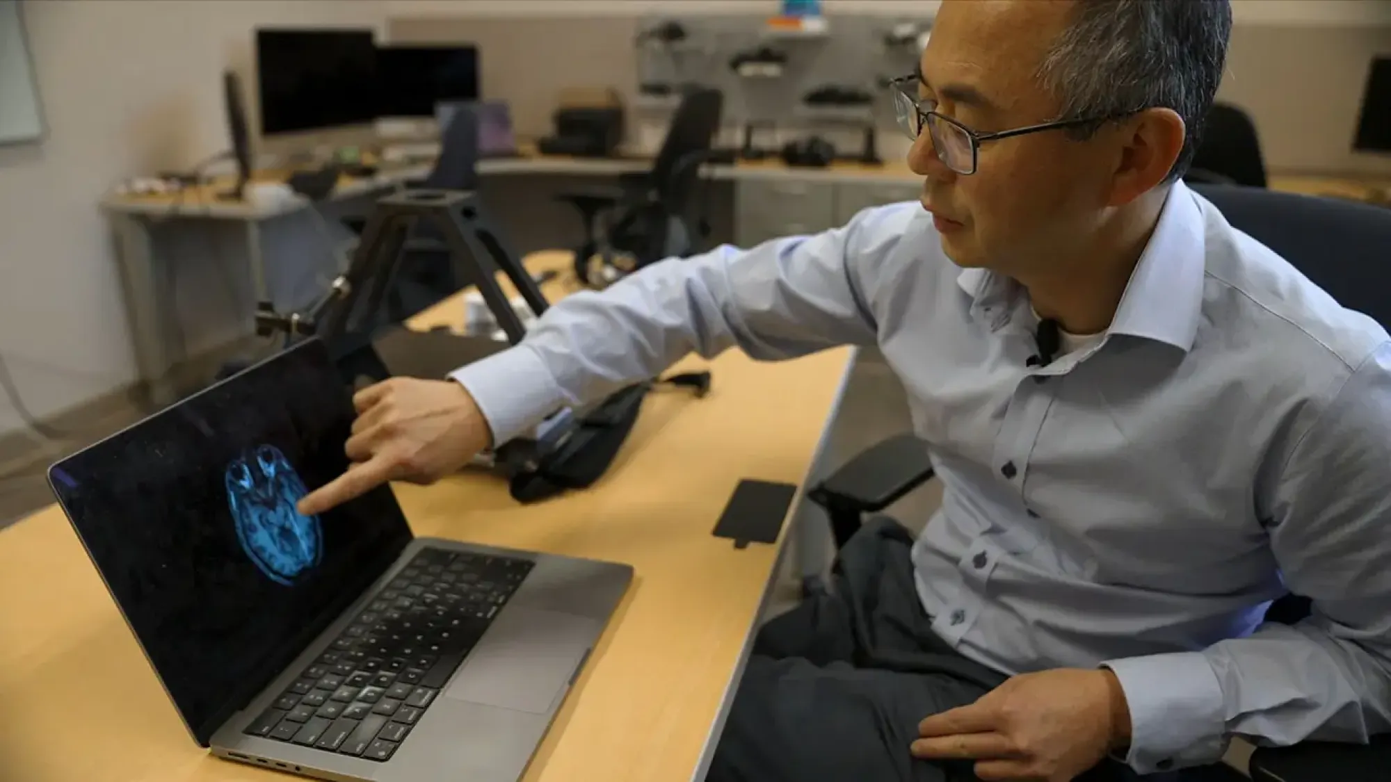 Man in office pointing at laptop screen showing blue brain scan.