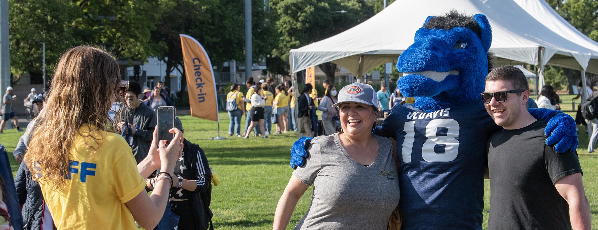Two adults pose with large blue mascot at a sunny park festival (photograph)