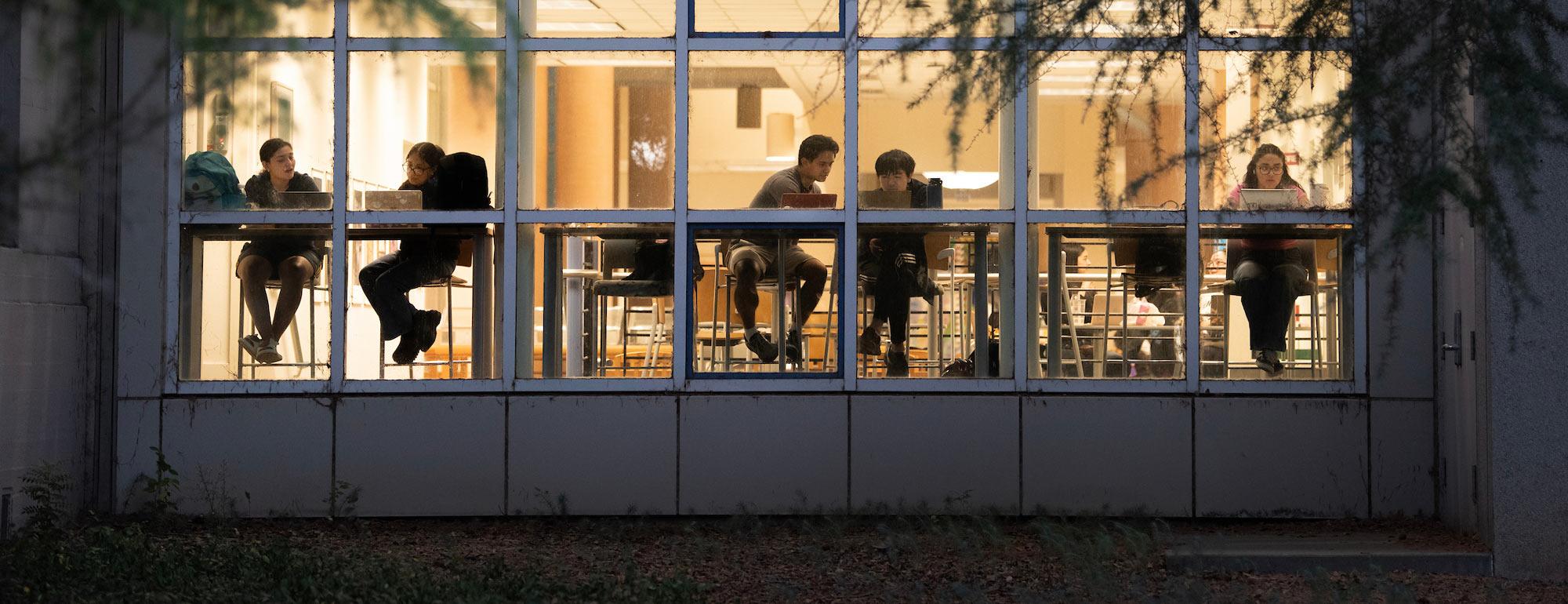 Warm-lit classroom seen through large windows at dusk, people silhouetted