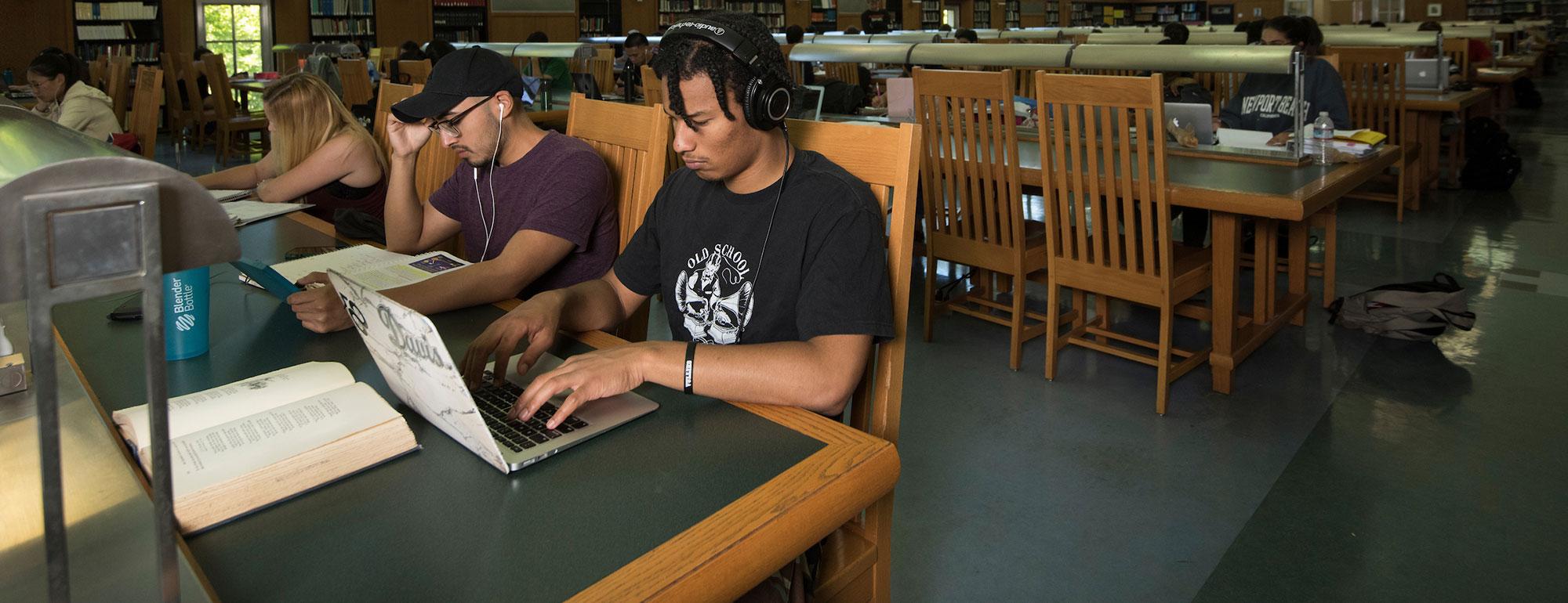 Students studying at green library table; one wearing headphones using a laptop.