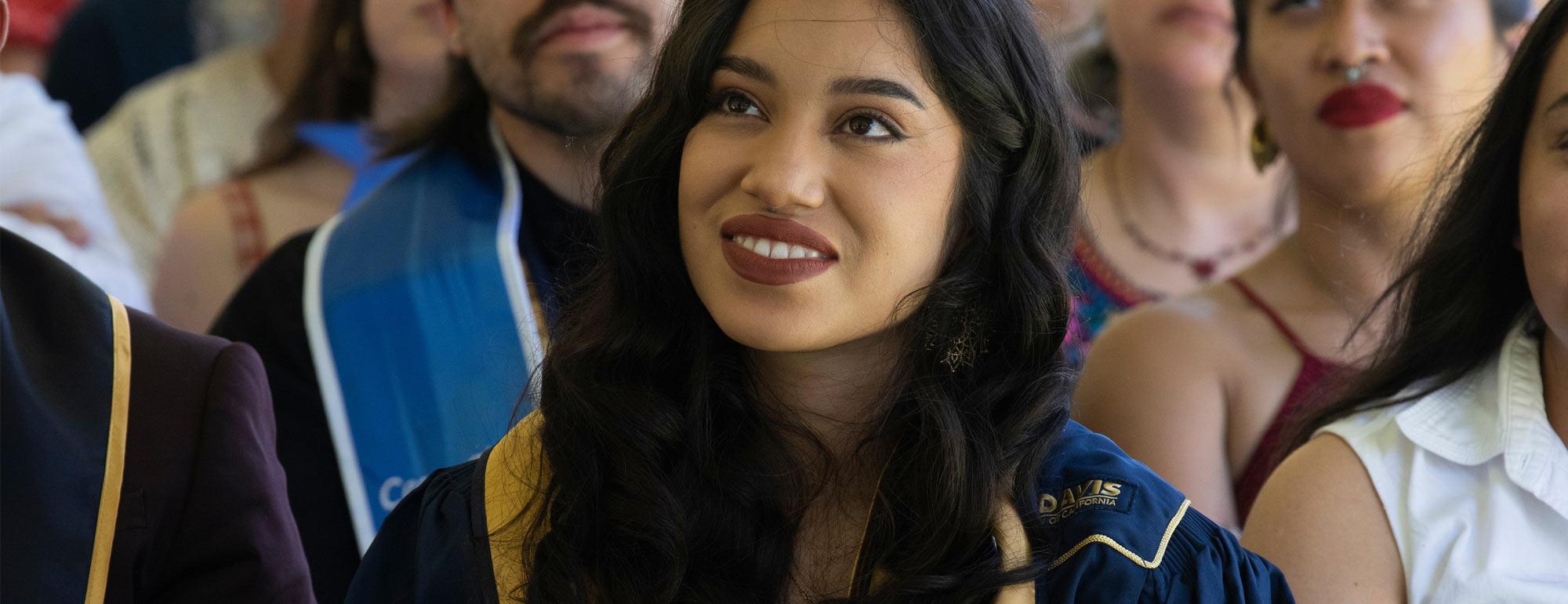 Smiling young woman in navy graduation gown and hood, seated among peers