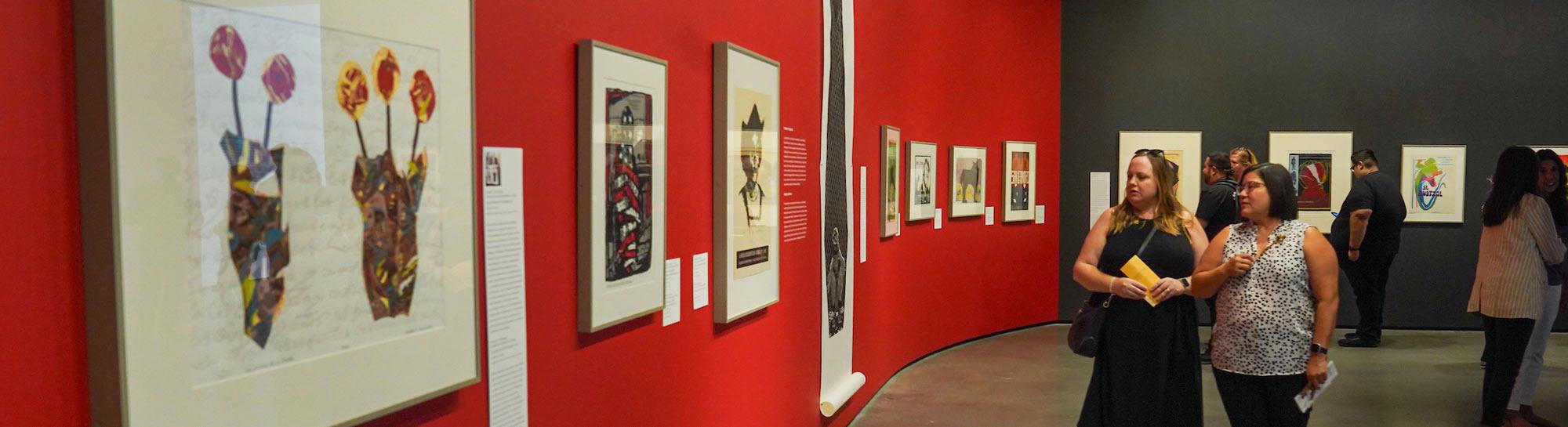 Visitors viewing framed artworks on red and dark gray gallery walls