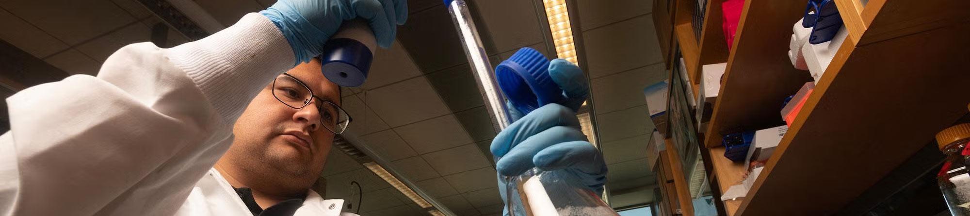 Lab worker in white coat and blue gloves reaching for a blue-capped bottle on shelf