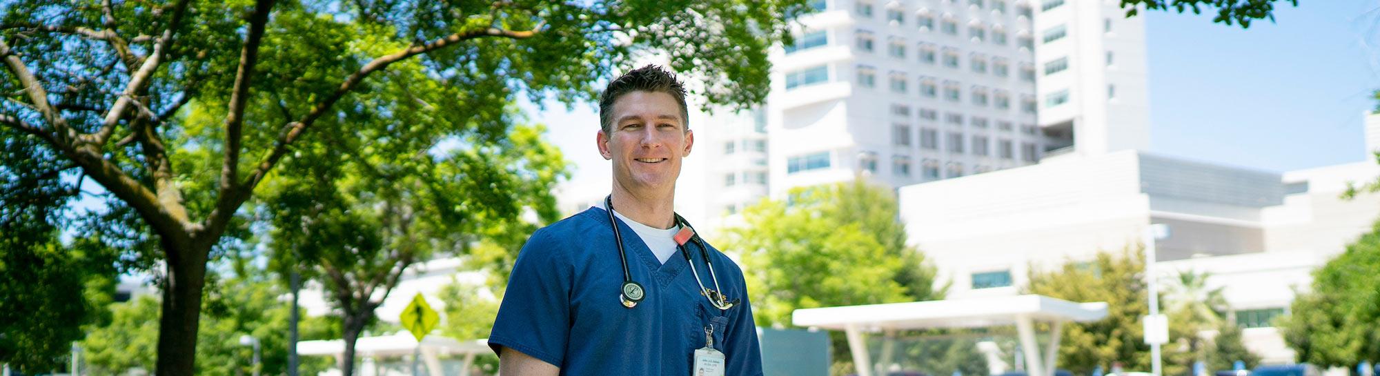 Young male medical professional in blue scrubs with stethoscope outdoors by hospital (photo)