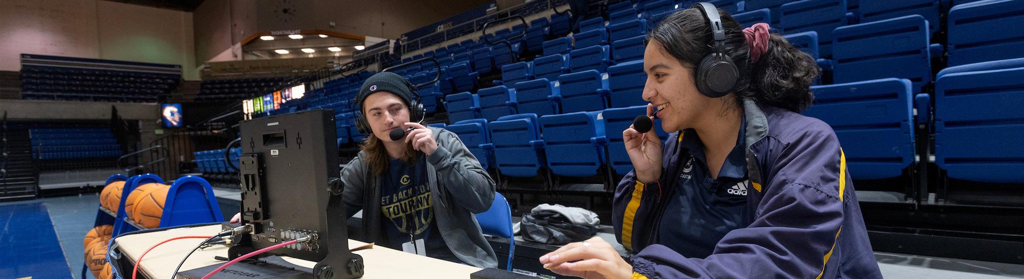 Two women broadcasters wearing headsets at a courtside table with laptop, empty blue bleachers