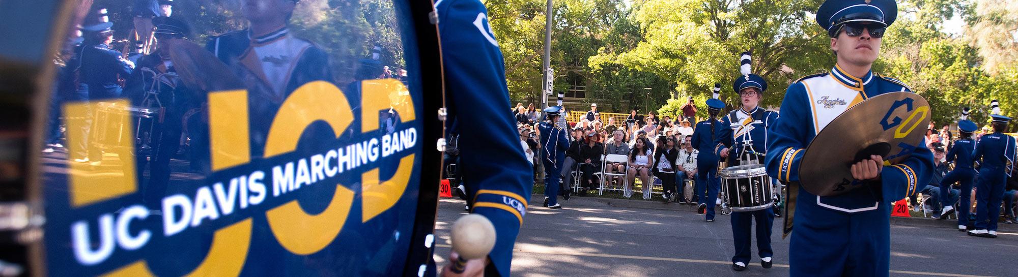 UC Davis marching band member in blue uniform playing trumpet beside banner at parade