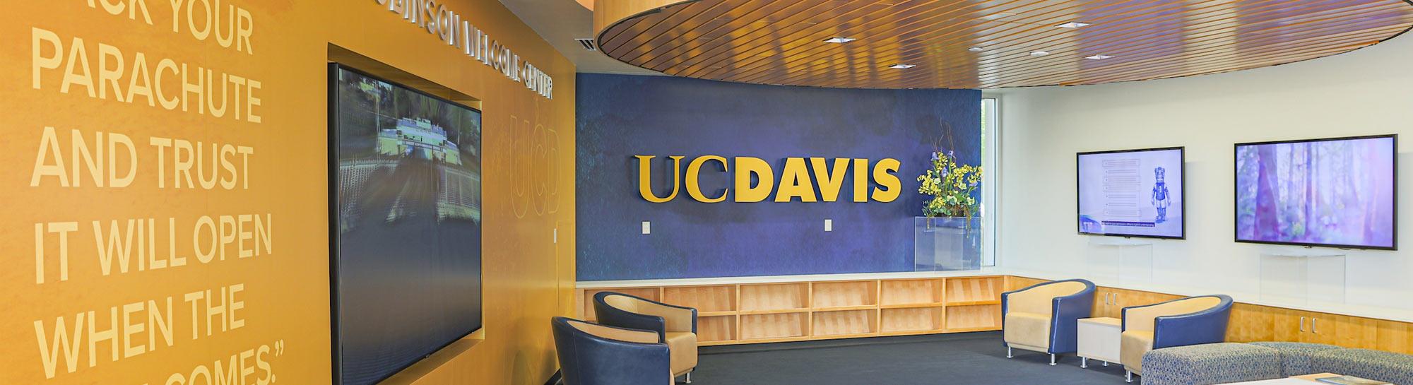 Campus lobby with large blue-and-gold UC Davis logo, wooden benches and windowed greenery