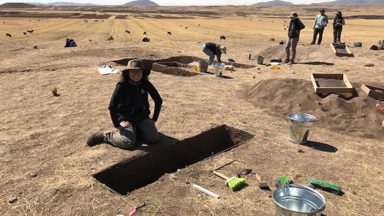 A student wearing a sunhat kneels in front of a trench on a sandy landscape. They are surrounded by tools, a team, and mountains