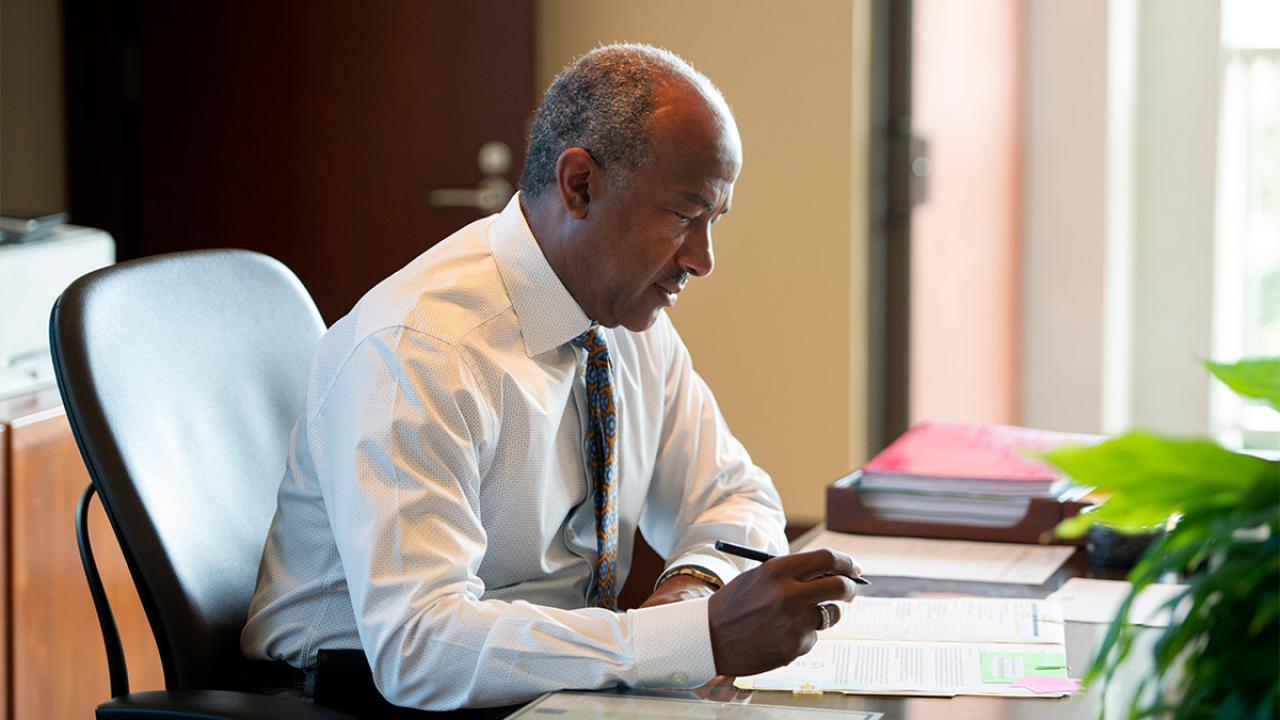 Chancellor Gary S. May sitting at a desk, writing.