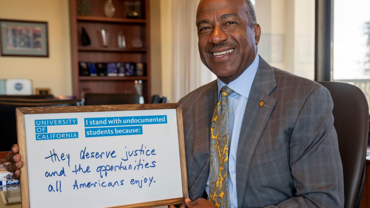 Chancellor Gary S. May, smiling, in his office, with DACA signboard.