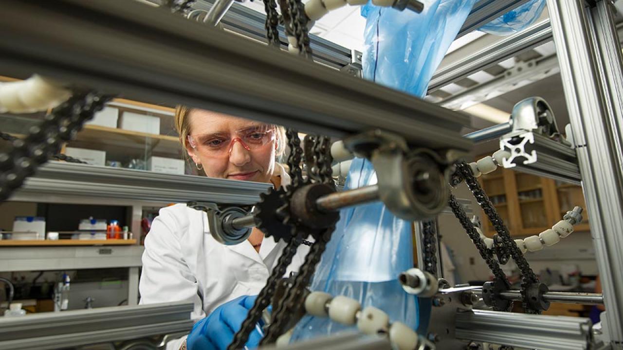 Woman in lab coat and goggles adjusts tabletop machine made of gears, chains and metal bars