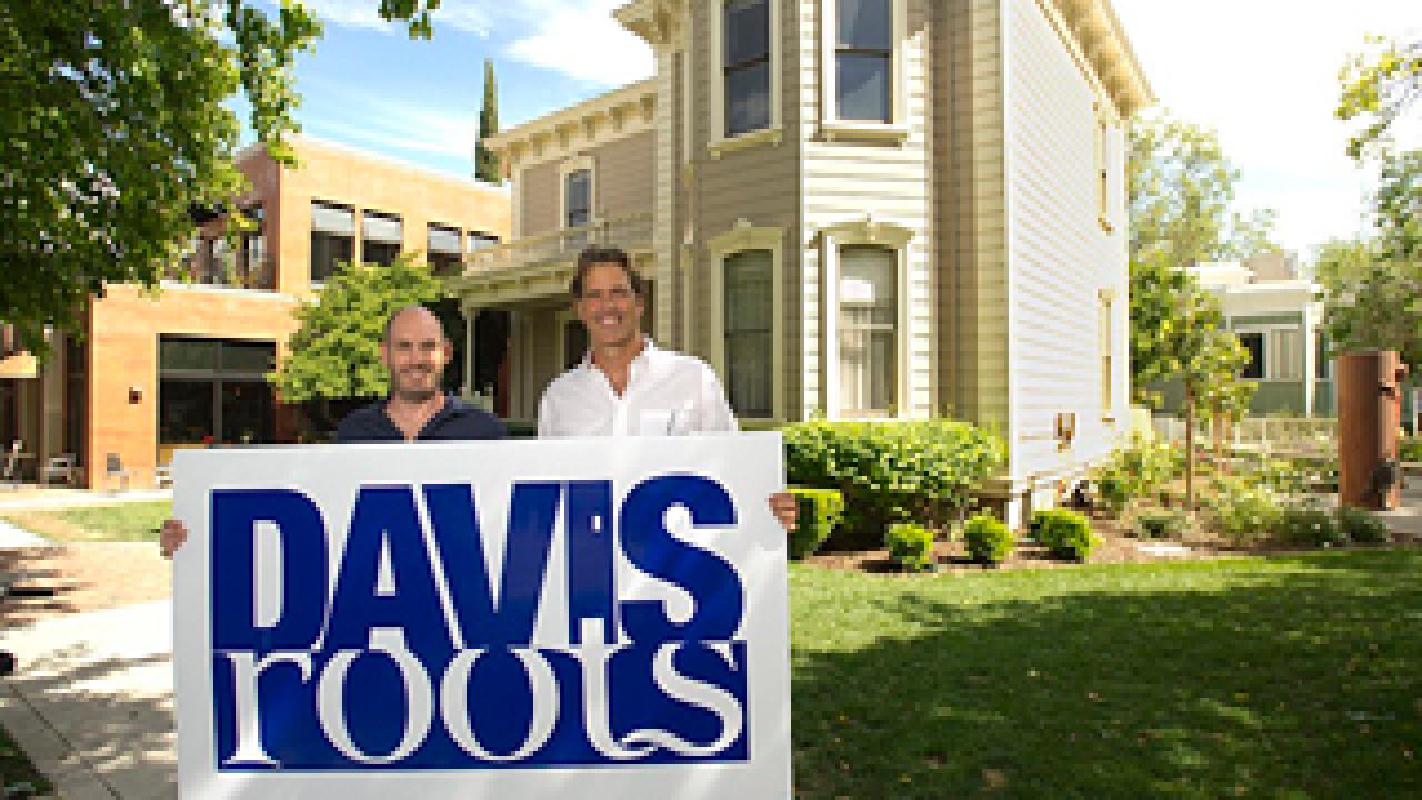 Two men standing behind a Davis Roots sign with a old mansion in the background