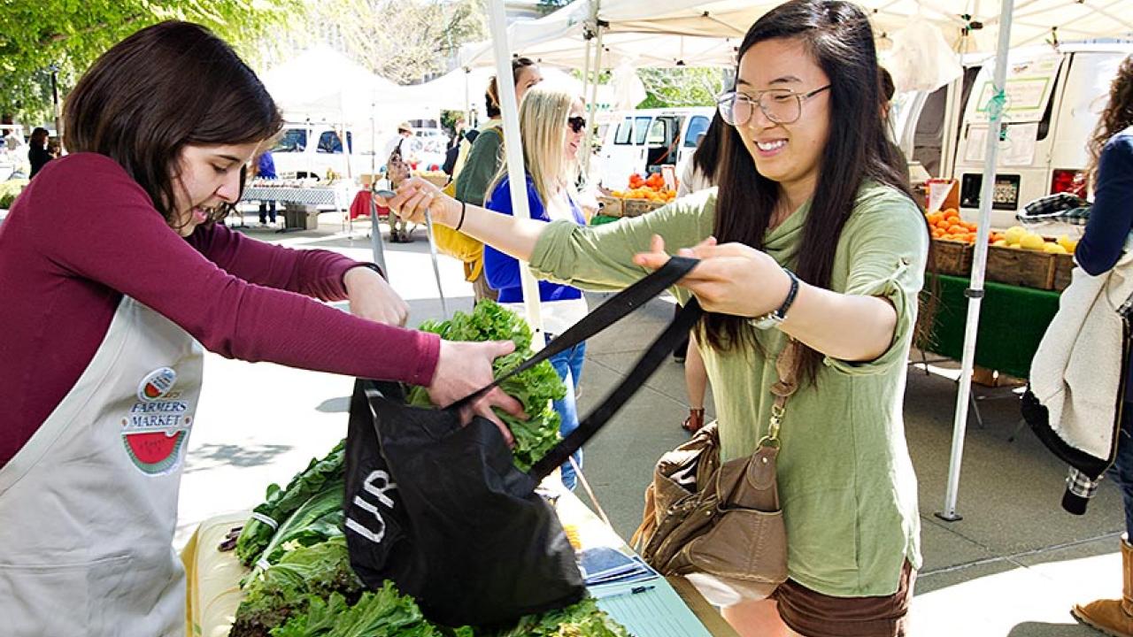 Woman places bunch of leaf lettuce in other woman's cloth shopping bag