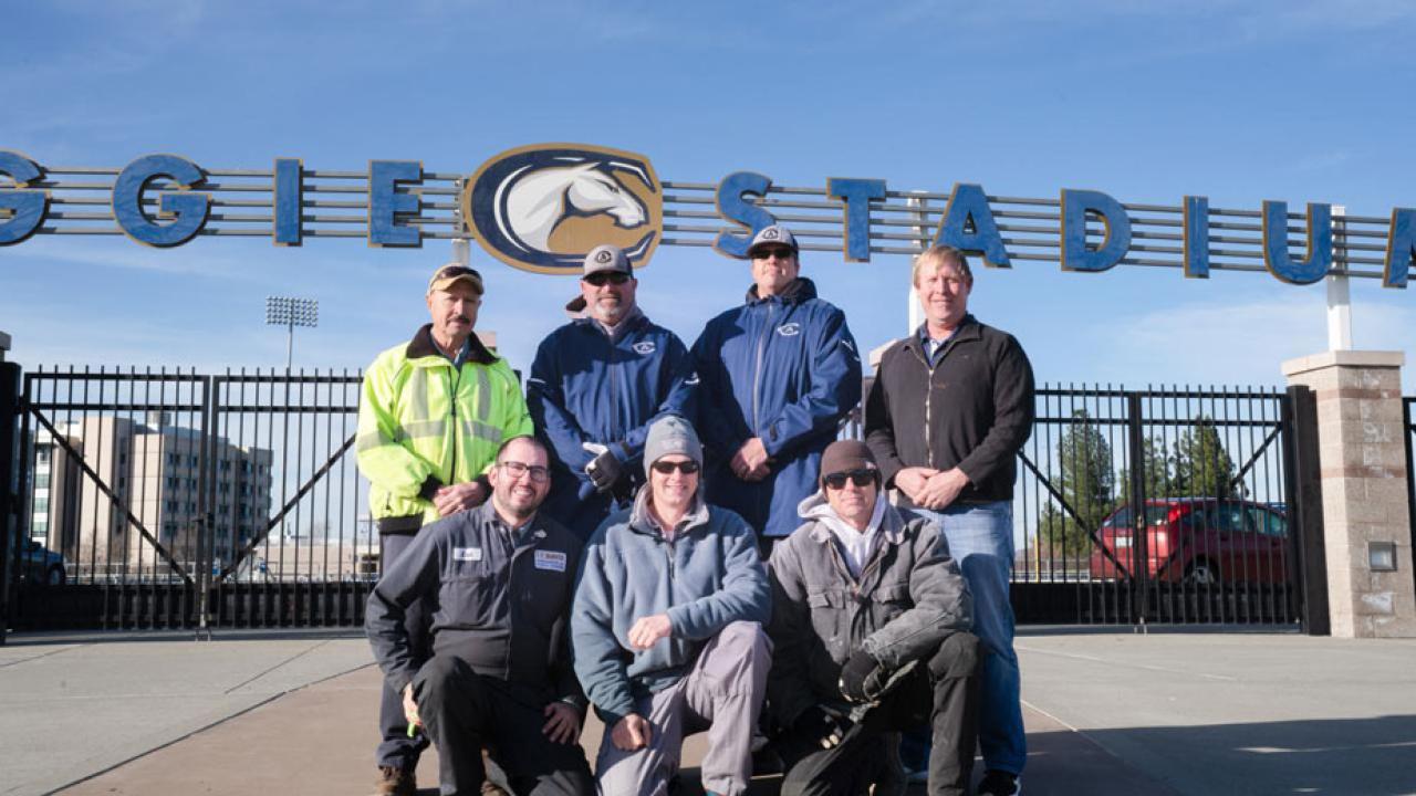 Seven men pose in front of Aggie Stadium gate and sign.