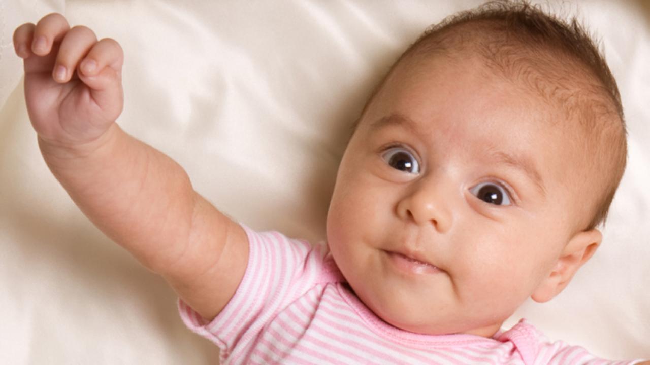 Infant girl with brown hair and brown eyes lies on her back,  reaching out one arm.