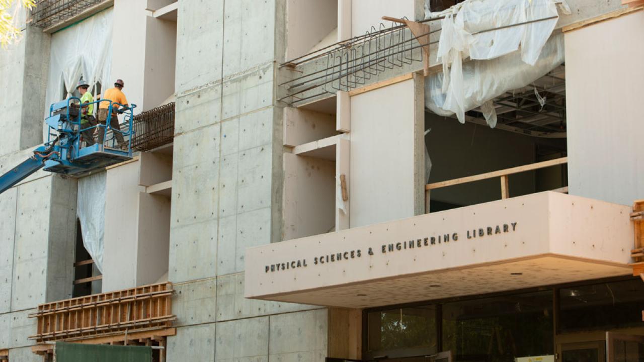 Workers in lift, on exterior of concrete building, with library's sign and front door in foreground.