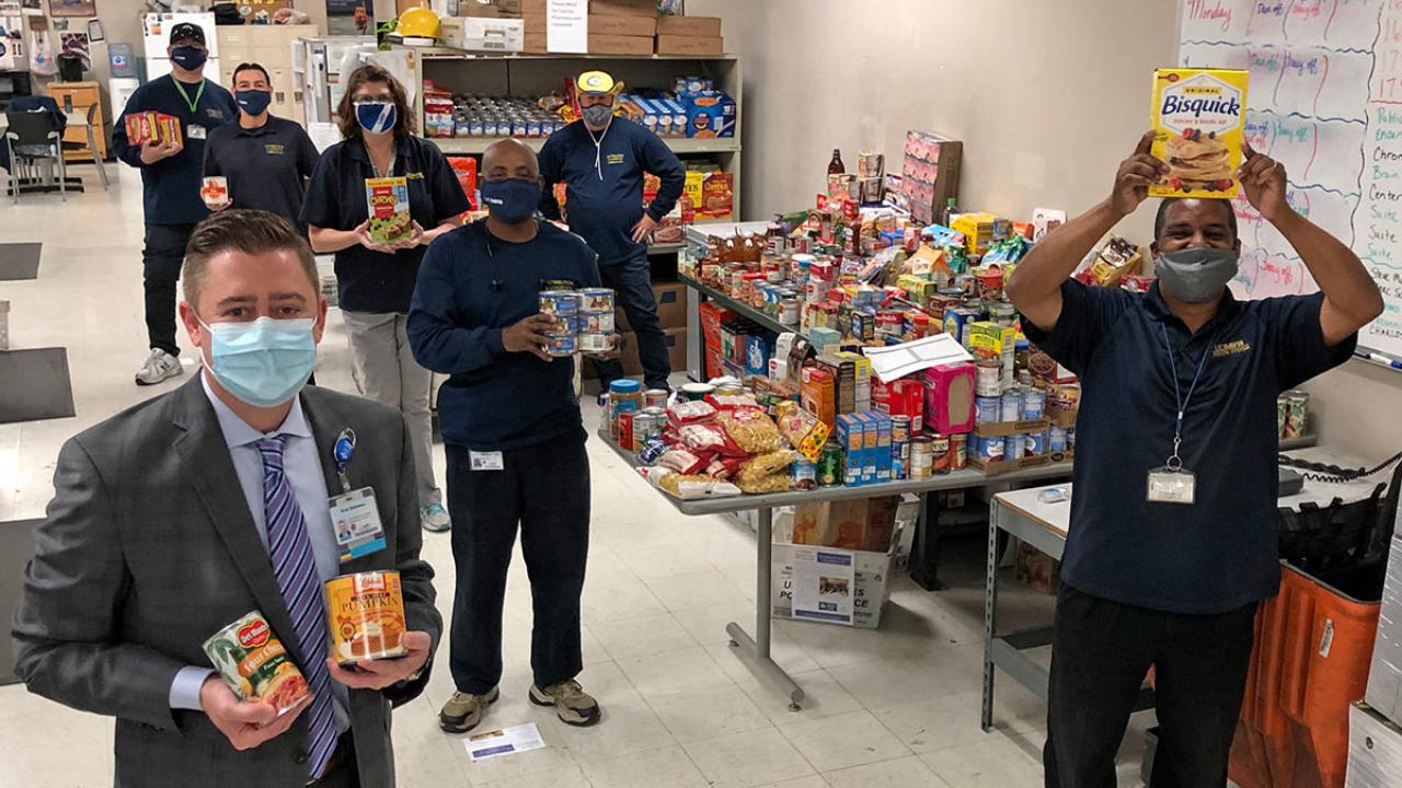 Mail Services crew and UC Davis Health executive hold food items, while standing around tables filled with food donations.