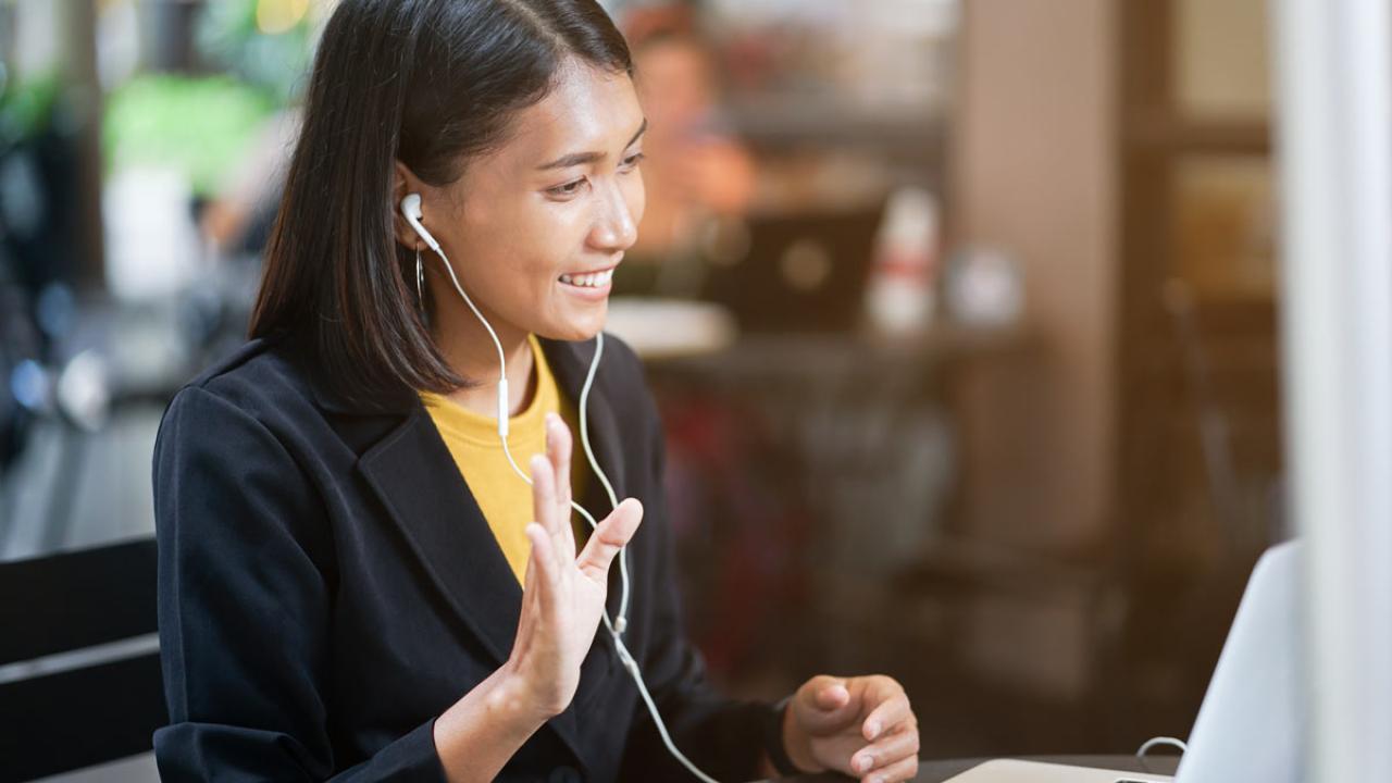 Woman, with earbuds, waves to computer screen, during a virtual interview.