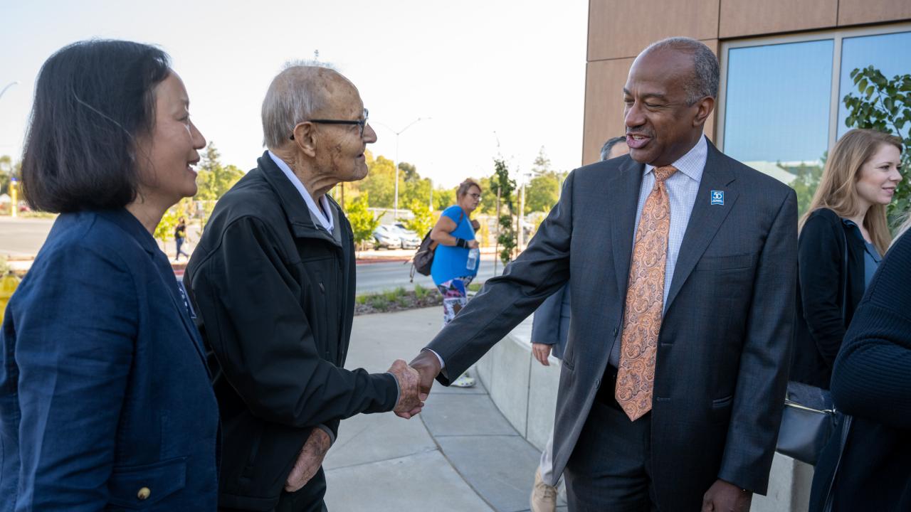 Elderly man with cane shakes hands, smiling with a suited official outdoors