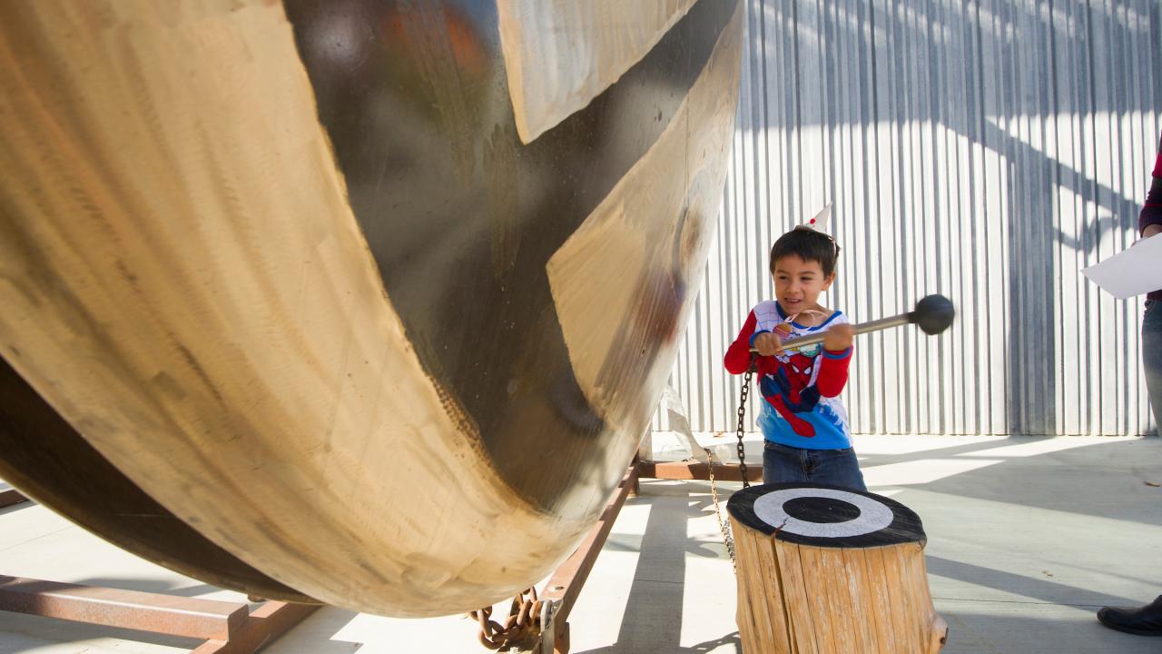 Child in superhero costume hitting a large gong sculpture 