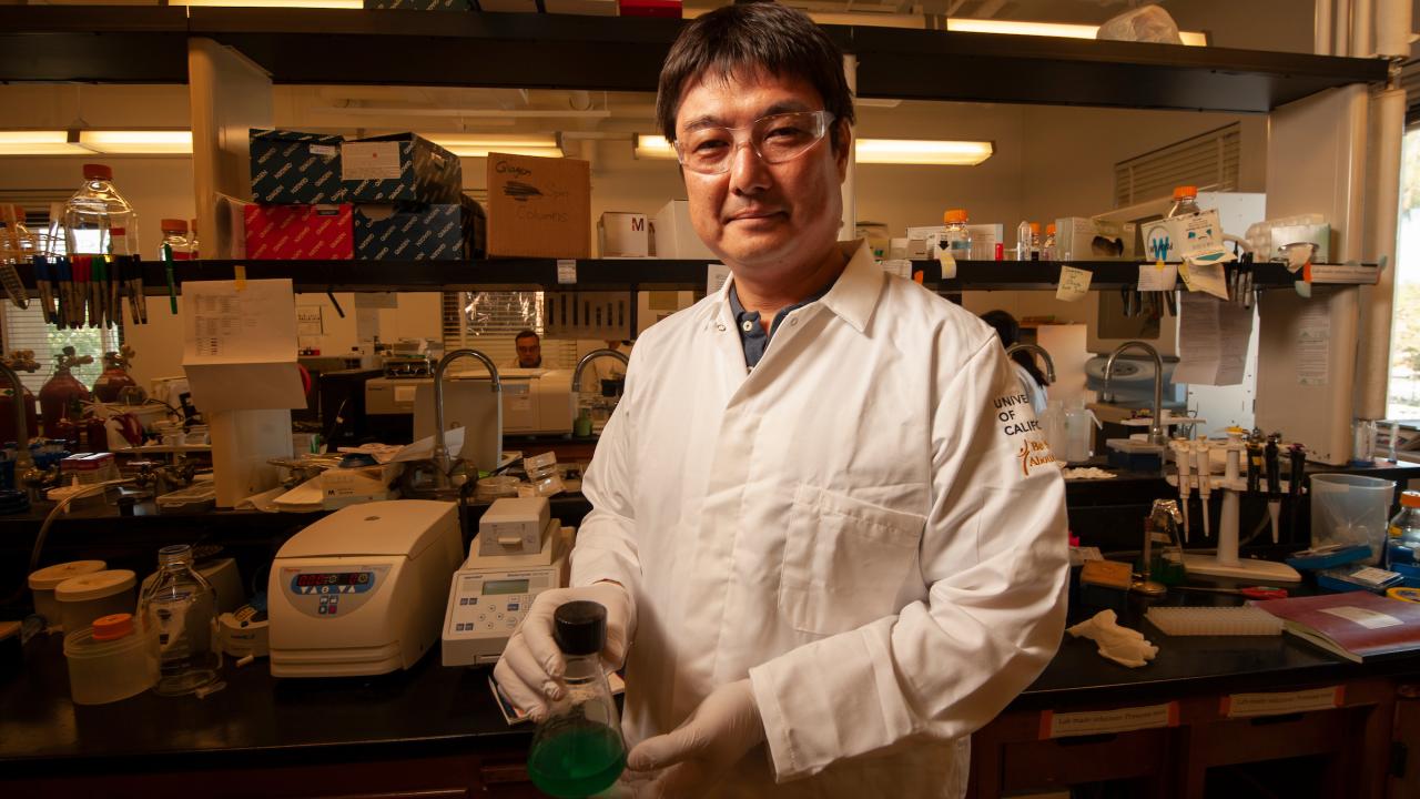 An Asian man with black hair wearing a white lab coat, gloves and safety glasses stands in front of a laboratory bench. 