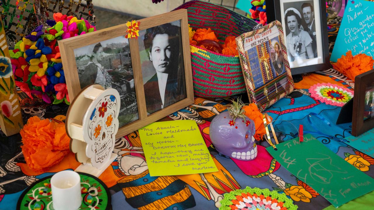 Framed photos and sculptures of sugar skulls sit atop an altar with a multicolor table cloth in an indoor setting.