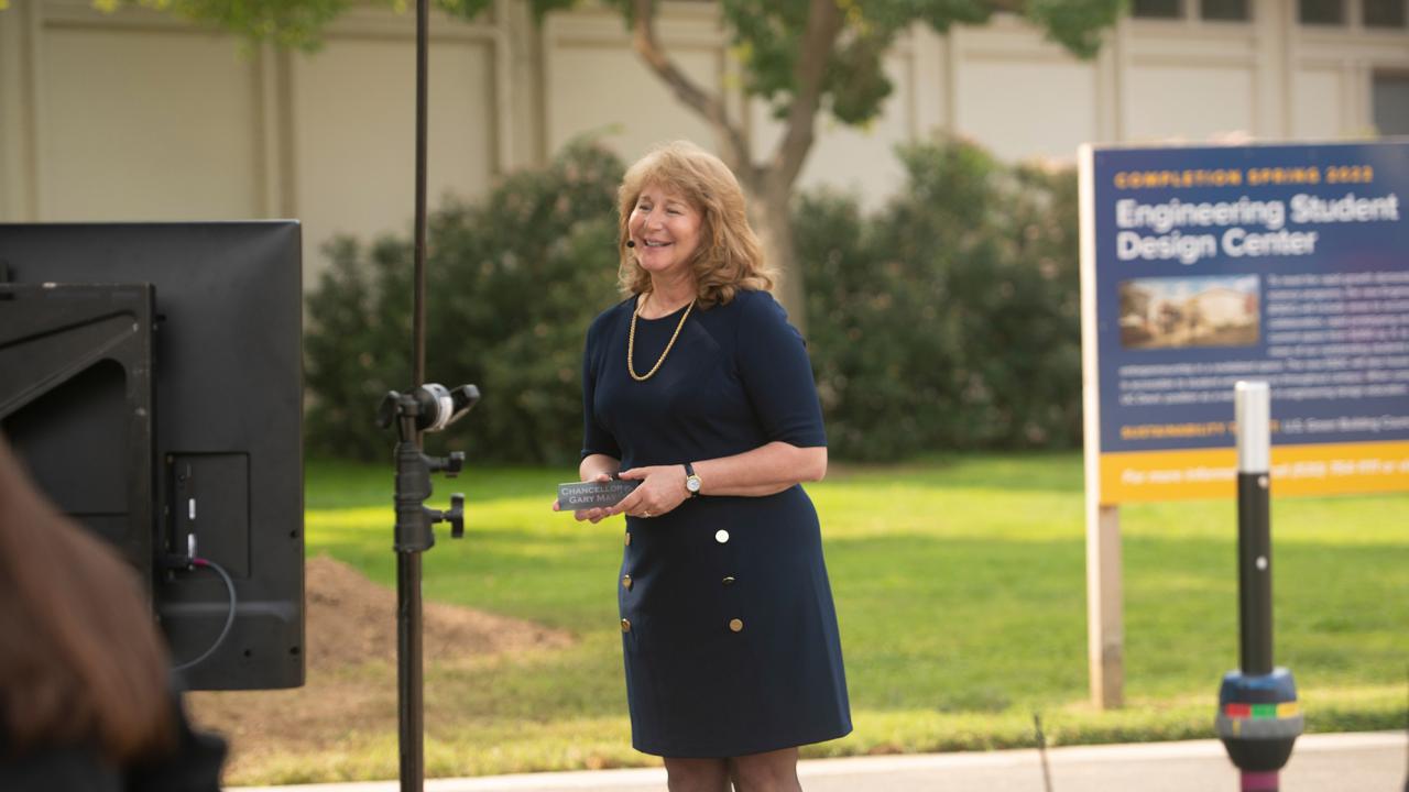 A blonde woman in a blue dress outdoors. To the left is the back of a video monitor and on the right is a blue sign slightly out of focus. 