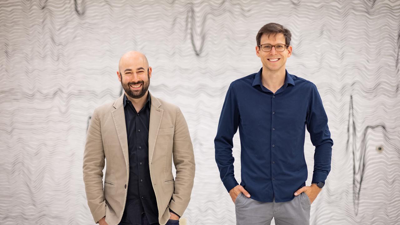 Two smiling men face the camera with hands in pockets. In the background and waveforms of brainwaves. 