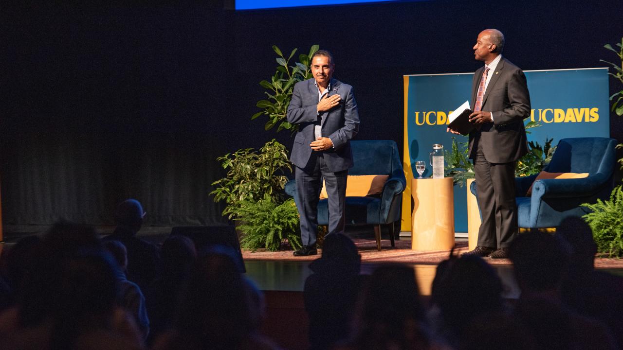 An audience inside a theater gives a standing ovation to a man on the theater stage, his hand over his heart in appreciation. Standing next to them is Chancellor Gary S May. 