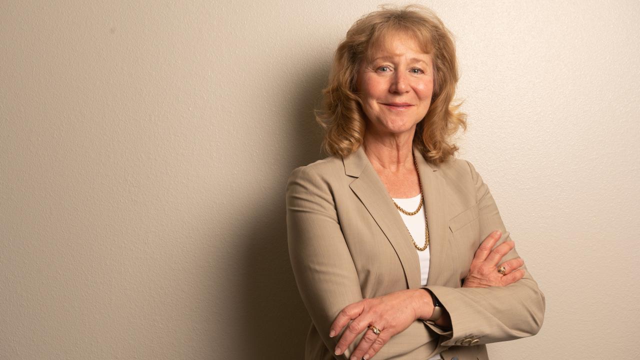 A photo of Jennifer Curtis, dressed in a beige colored business suit with a white blouse and off-white background, standing with her arms crossed.