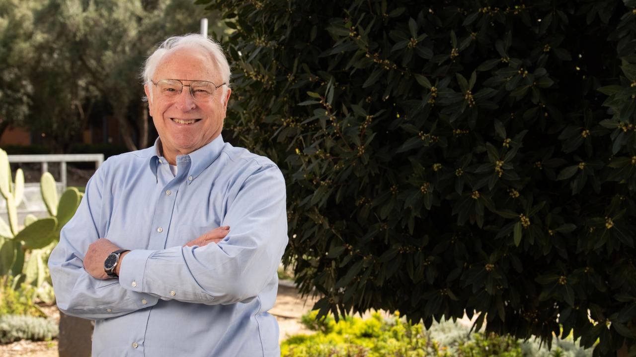 A man standing outside with his arms crossed against a button up shirt smiles at the camera at dusk.