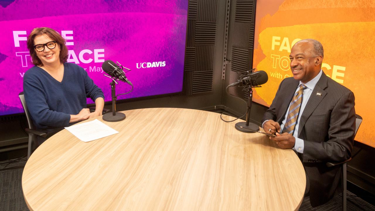 In a recording studio setting, a woman in a blue shirt and glasses sits across the table from Chancellor Gary S. May wearing a dark suit and tie. Microphones rest on the table between them. 