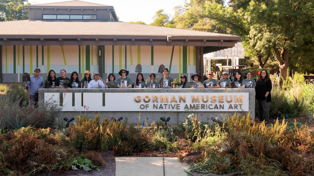 Group of people on a museum terrace behind a stone sign, mid-century building and gardens
