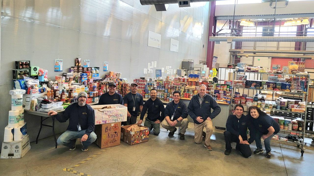 Eight people pose for photo in front of large pile of canned food and other supplies