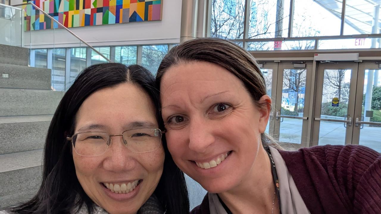 Two women smiling for a selfie in a bright atrium with colorful wall art and hanging glass orbs