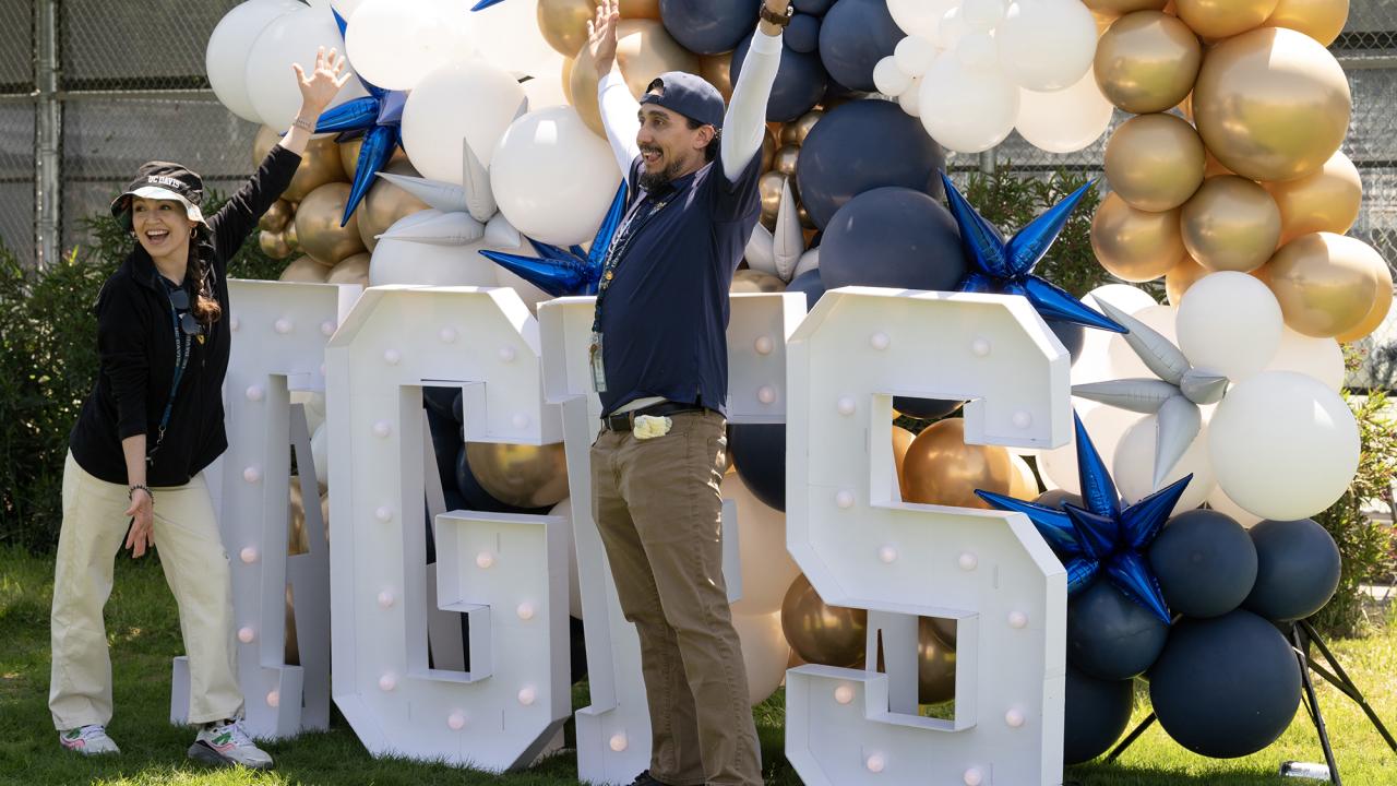 Three people celebrating by large white letters and gold, black, blue balloons outdoors